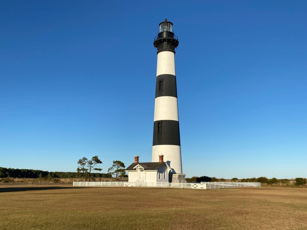 The horizontally banded black and white Bodie Island Lighthouse rises above the trees against a clear blue sky.