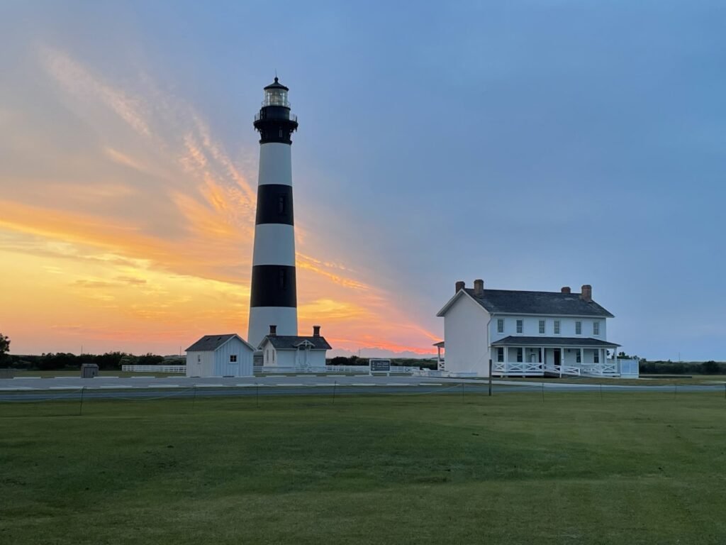 The black and white banded Bodie Island Lighthouse and its keepers' quarters lit by an orange sunrise.