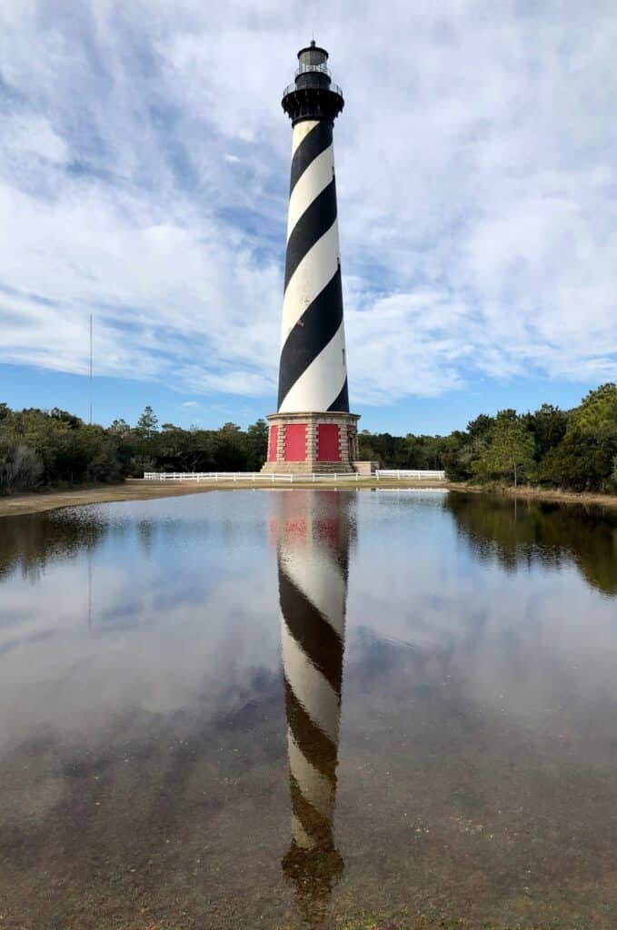 The black and white spiral-banded Cape Hatteras Lighthouse mirrored in standing floodwater at Buxton, framed by pines and blue sky.
