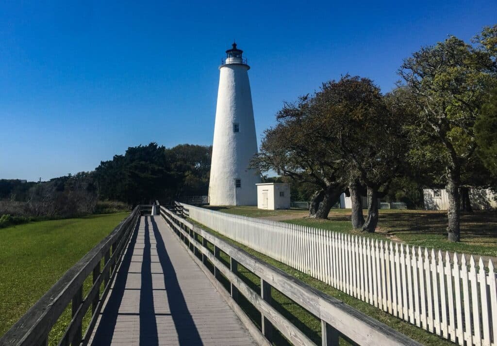 The short, whitewashed Ocracoke Lighthouse at the end of a white picket fence and boardwalk in Ocracoke Village.