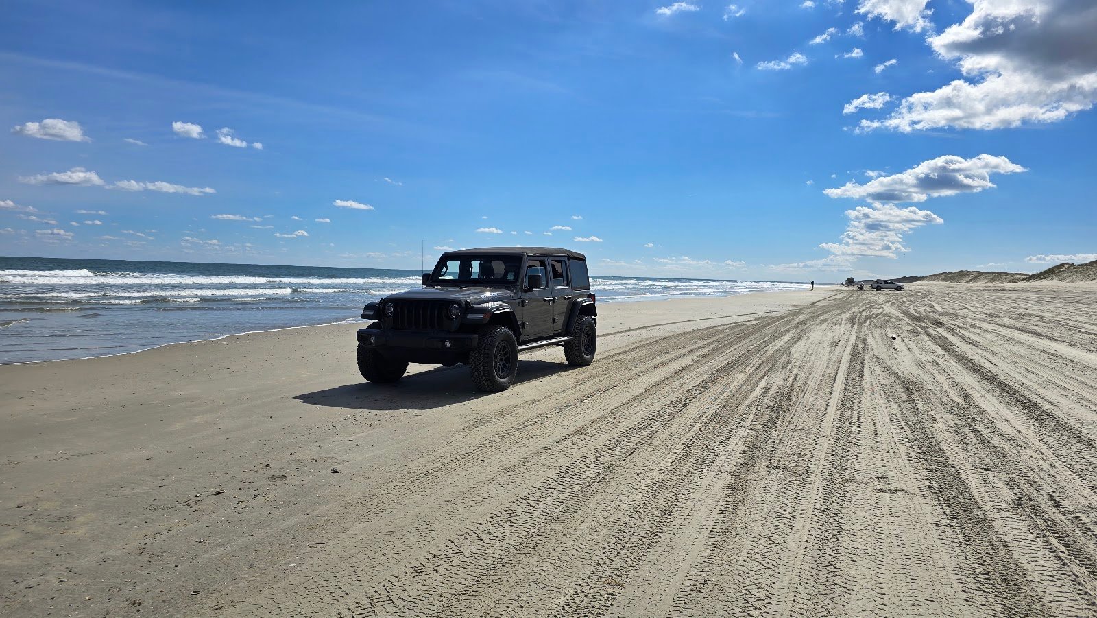 A Jeep Wrangler driving the open sand on the Outer Banks, tire tracks running down the beach with surf alongside