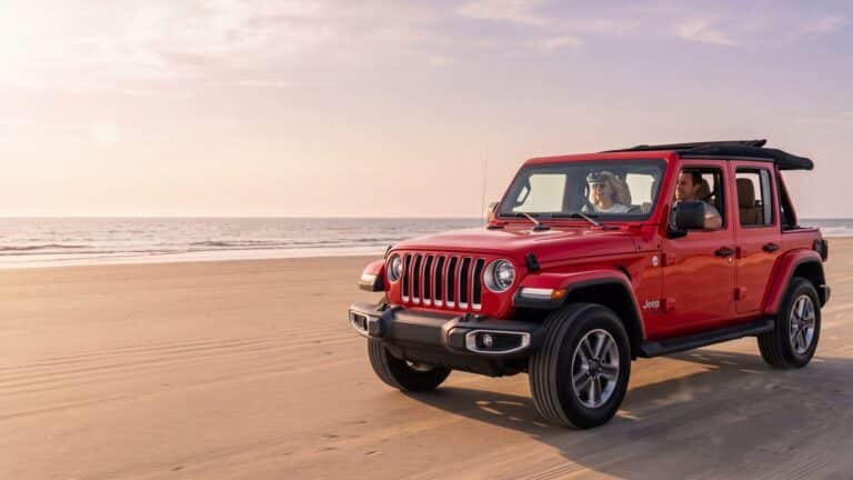 A red four-door Jeep Wrangler driving on hard sand with a couple inside, both seated and belted — Outer Banks, NC