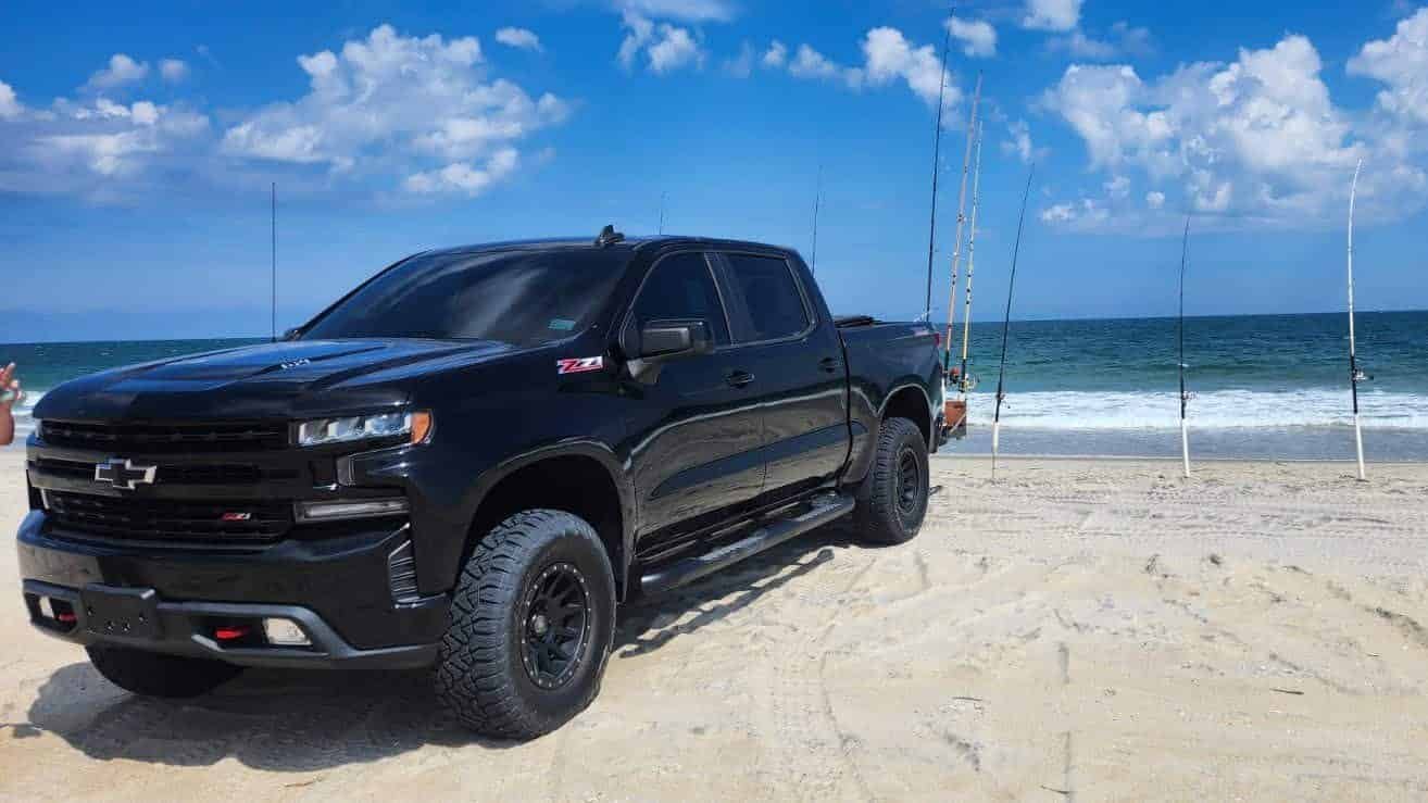 Real photo: Beach 4x4’s black Chevrolet Silverado Trail Boss parked on the beach with surf-fishing rods in the sand.