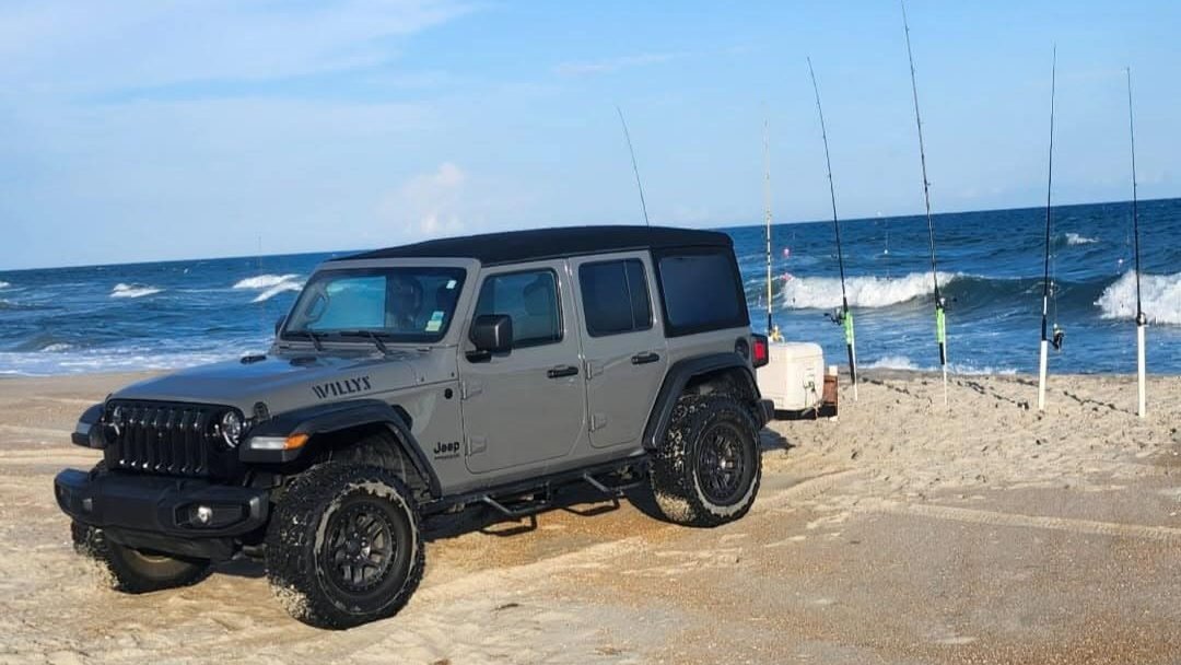 Real photo: a gray Jeep Wrangler Willys on the Carova beach with surf-fishing rods in the sand.